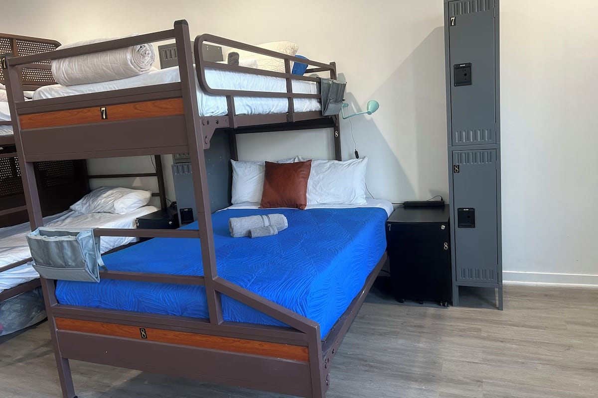 Ein Etagenbett mit blauer Matratze, einem Holz-Kopfstück und einer grauen Decke. Ein kleiner Nachttisch mit Lampe und Steckdose ist ebenfalls vorhanden.