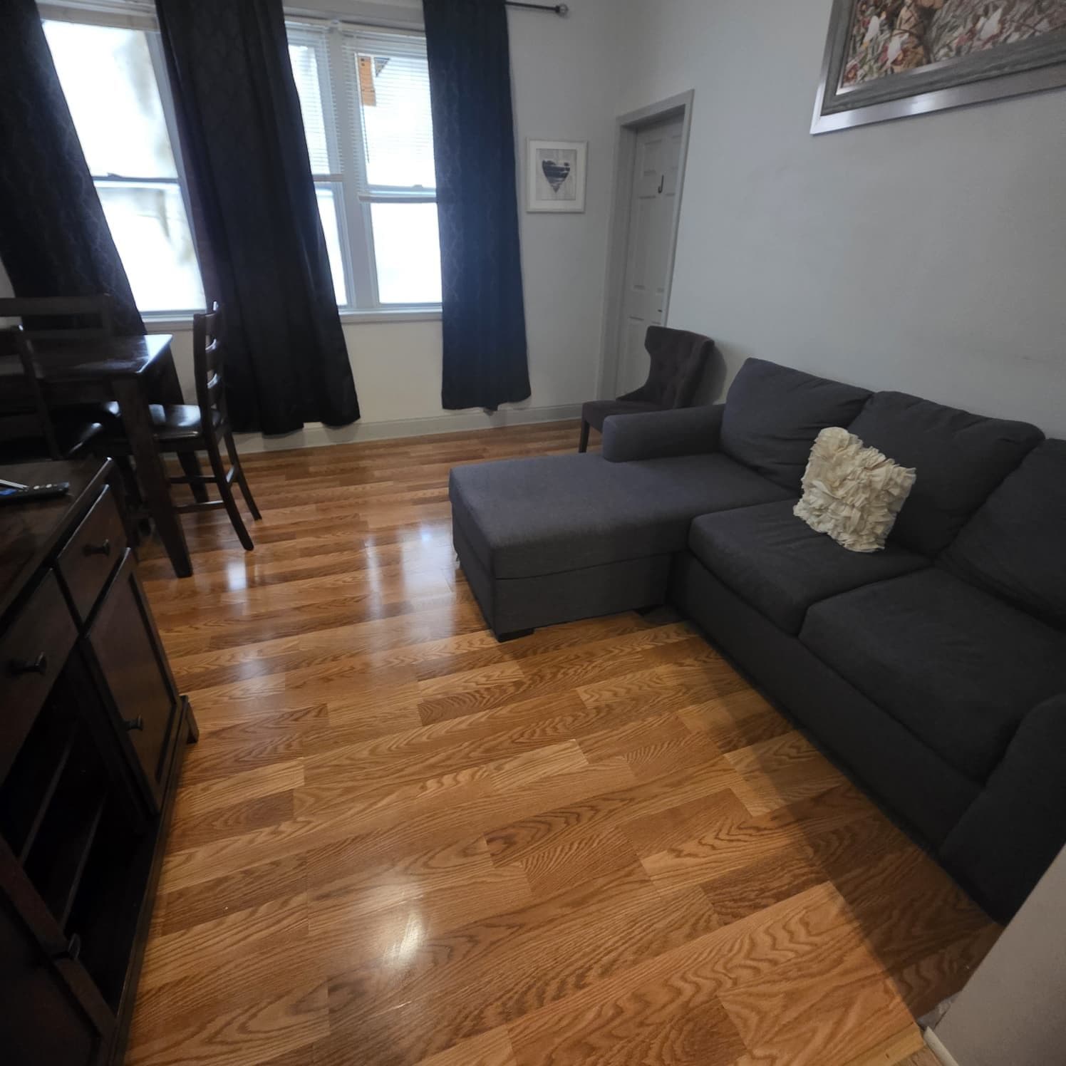 Una sala de estar con un sofá gris oscuro, piso de madera, una mesa de comedor con sillas y una ventana con cortinas.