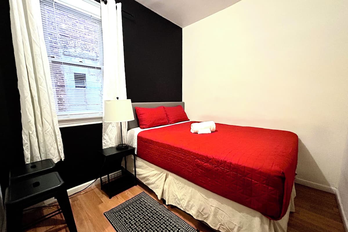 Ein Hotelzimmer mit Bett, Nachttisch, Lampe, Stuhl und einem Fenster mit Jalousien.