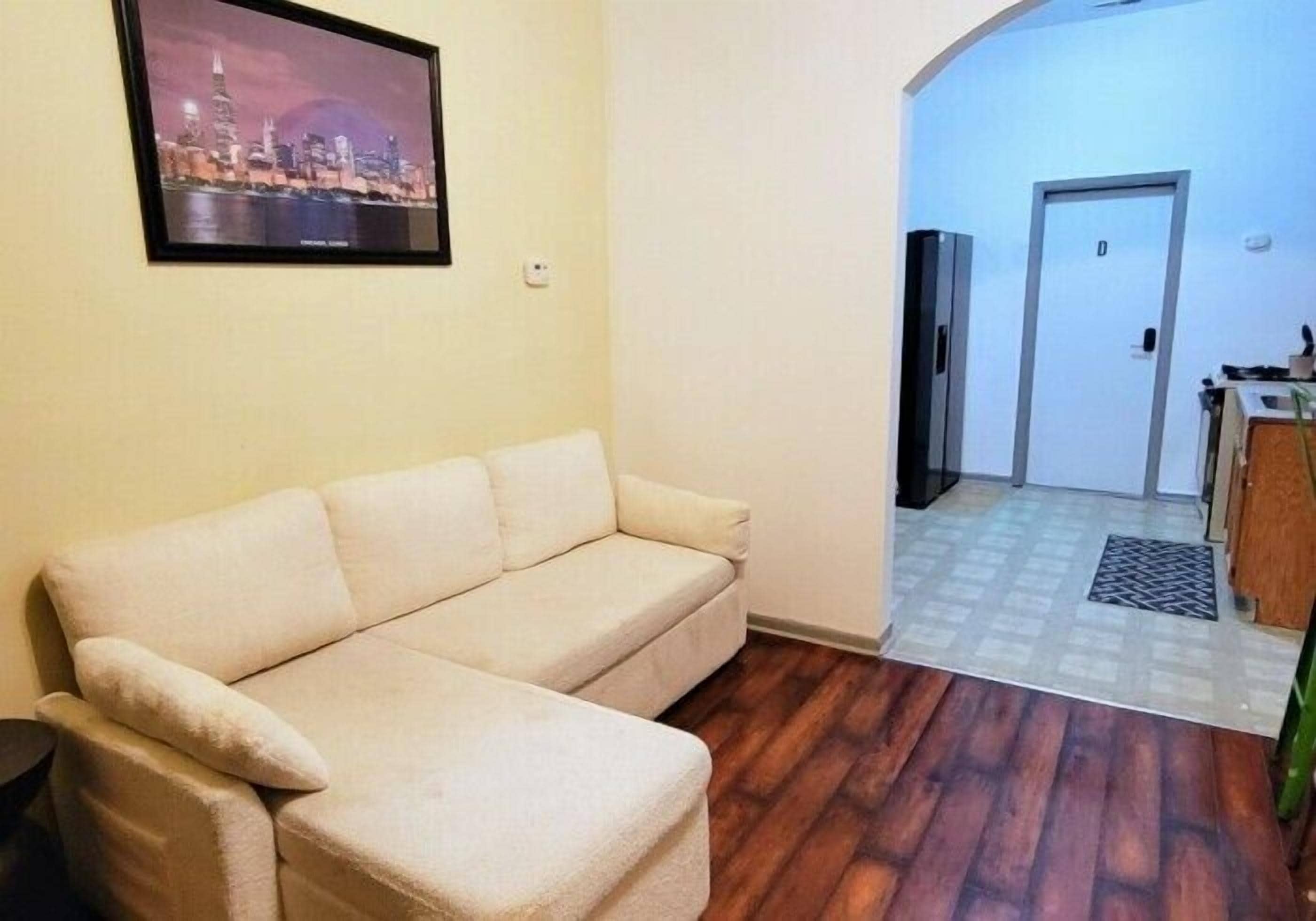 A living room with a sofa, a framed picture on the wall, a wooden floor, and a kitchen area visible through an archway.