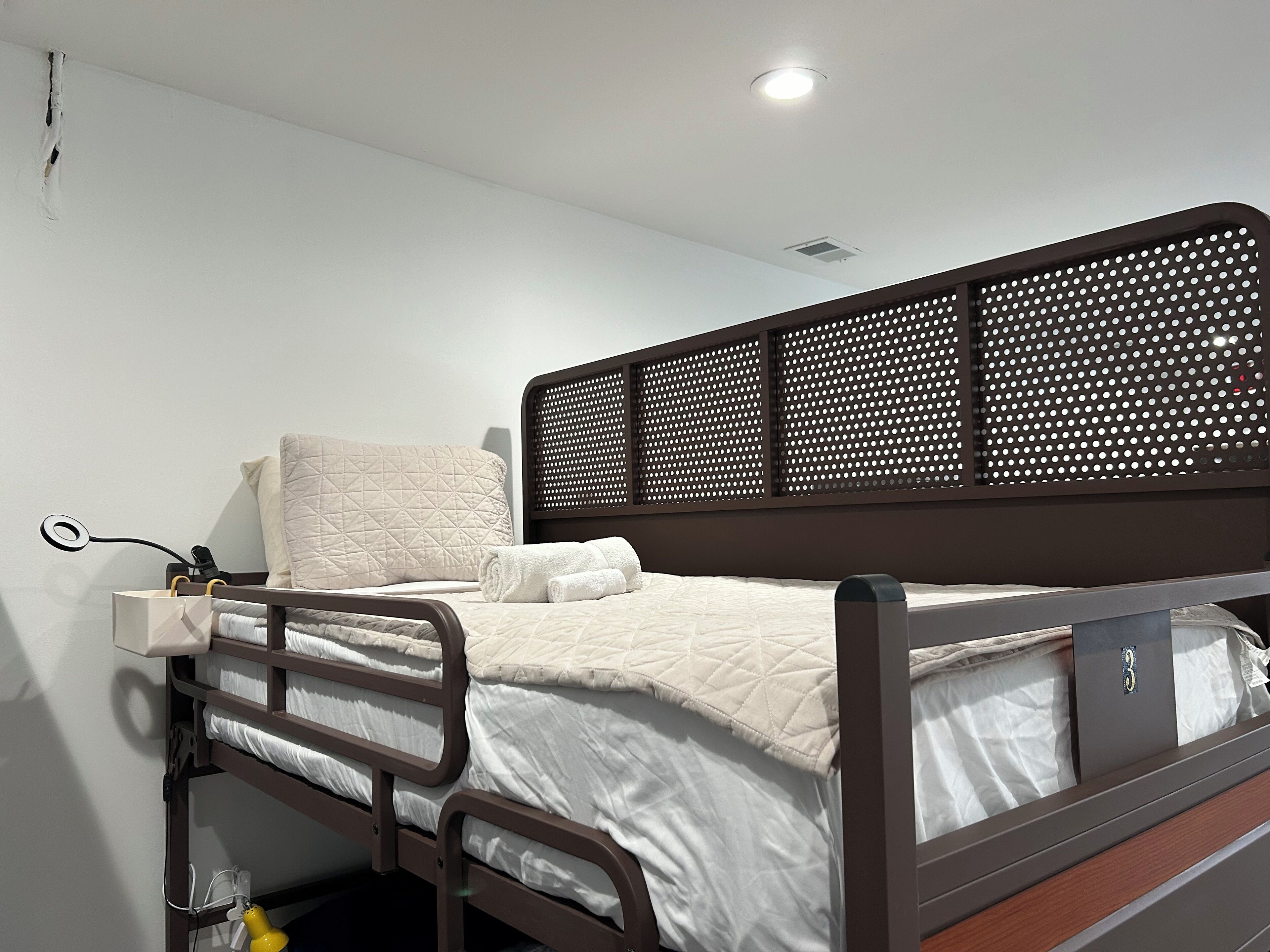Habitación individual con una cama de madera, una mesita de noche con lámpara y un enchufe en la pared.