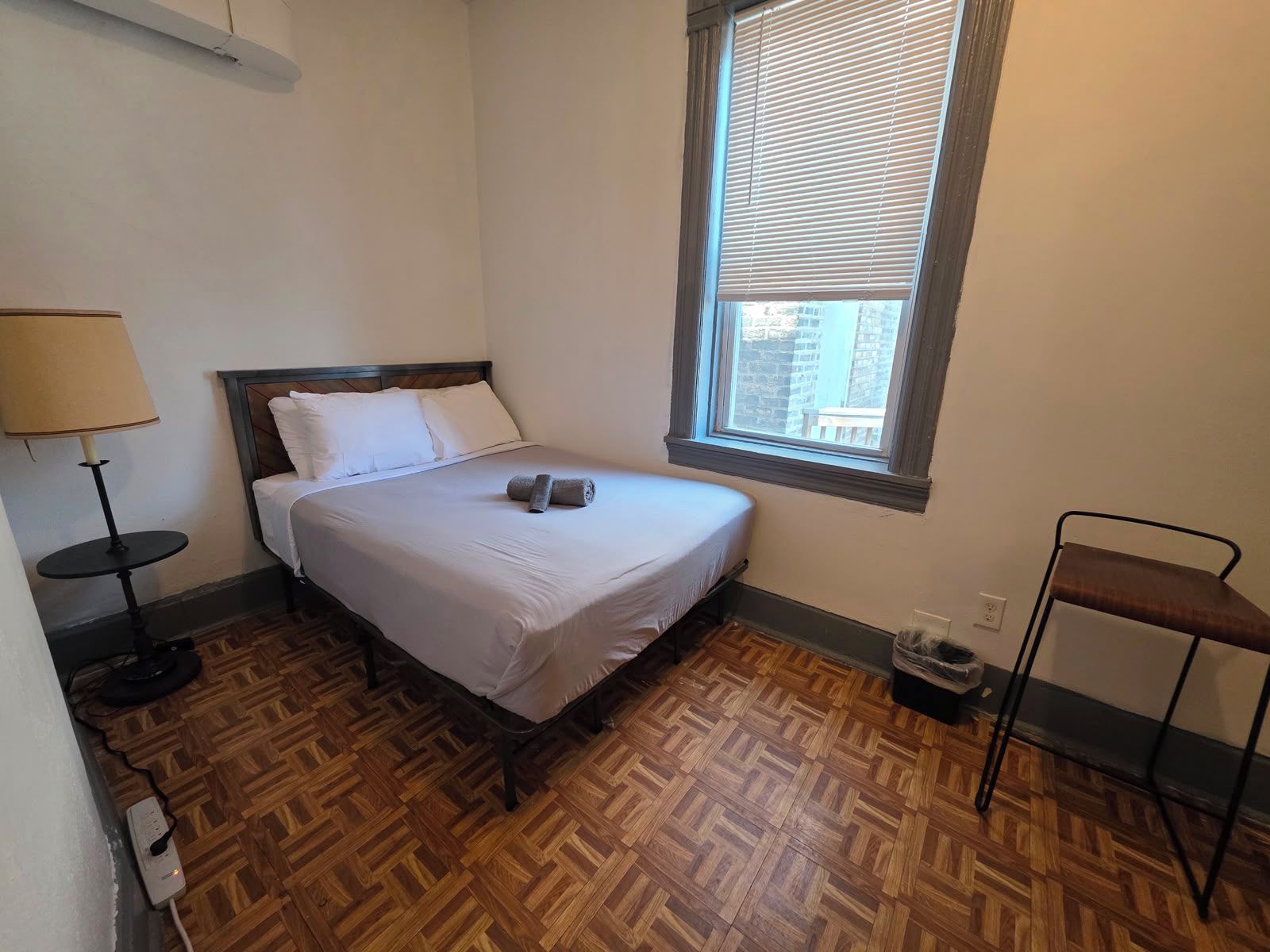 A small, neatly arranged bedroom with a bed, a bedside table, a lamp, a window with blinds, and a chair.