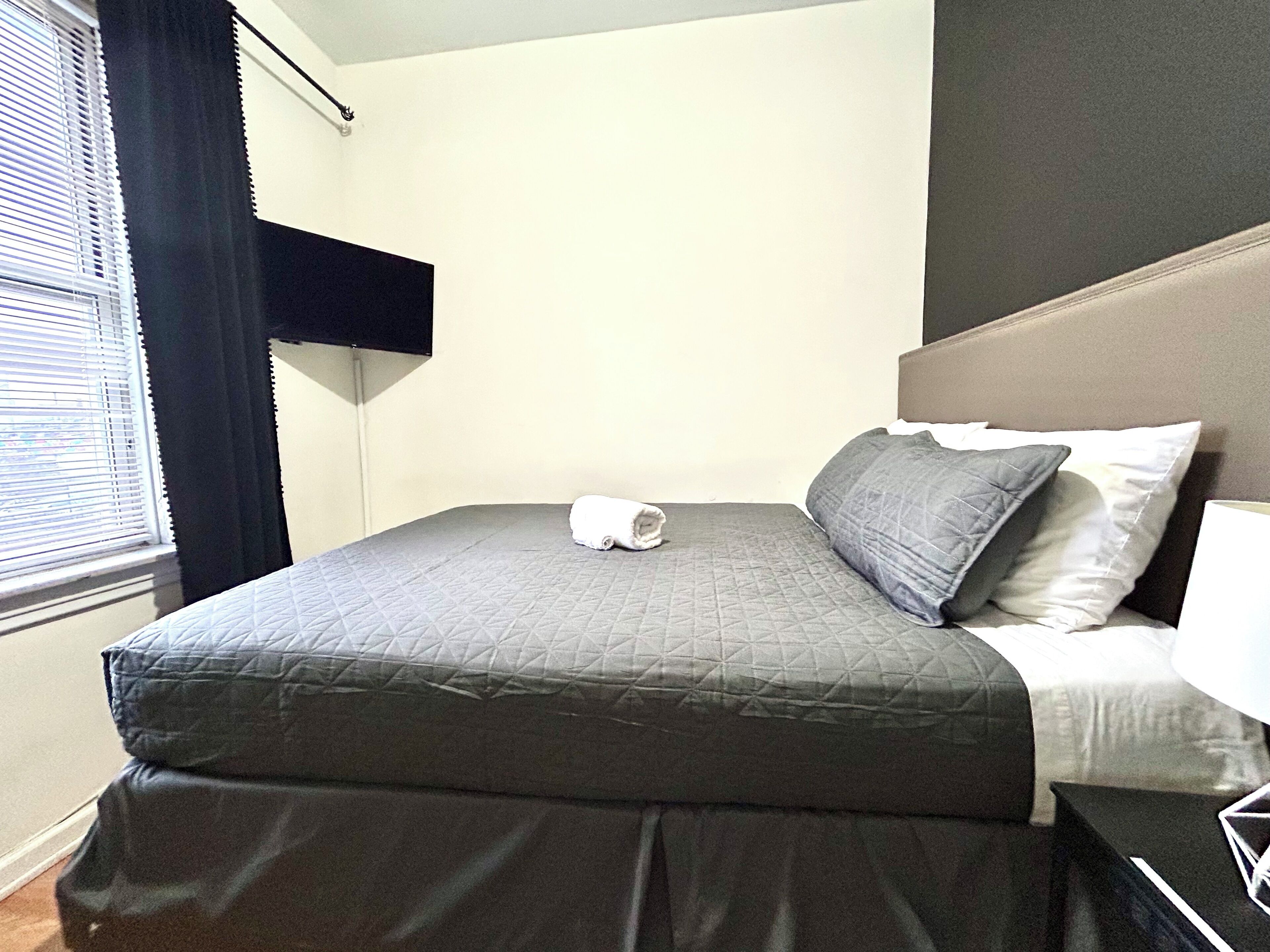 Una cama bien hecha con un edredón gris, una toalla enrollada y dos almohadas.