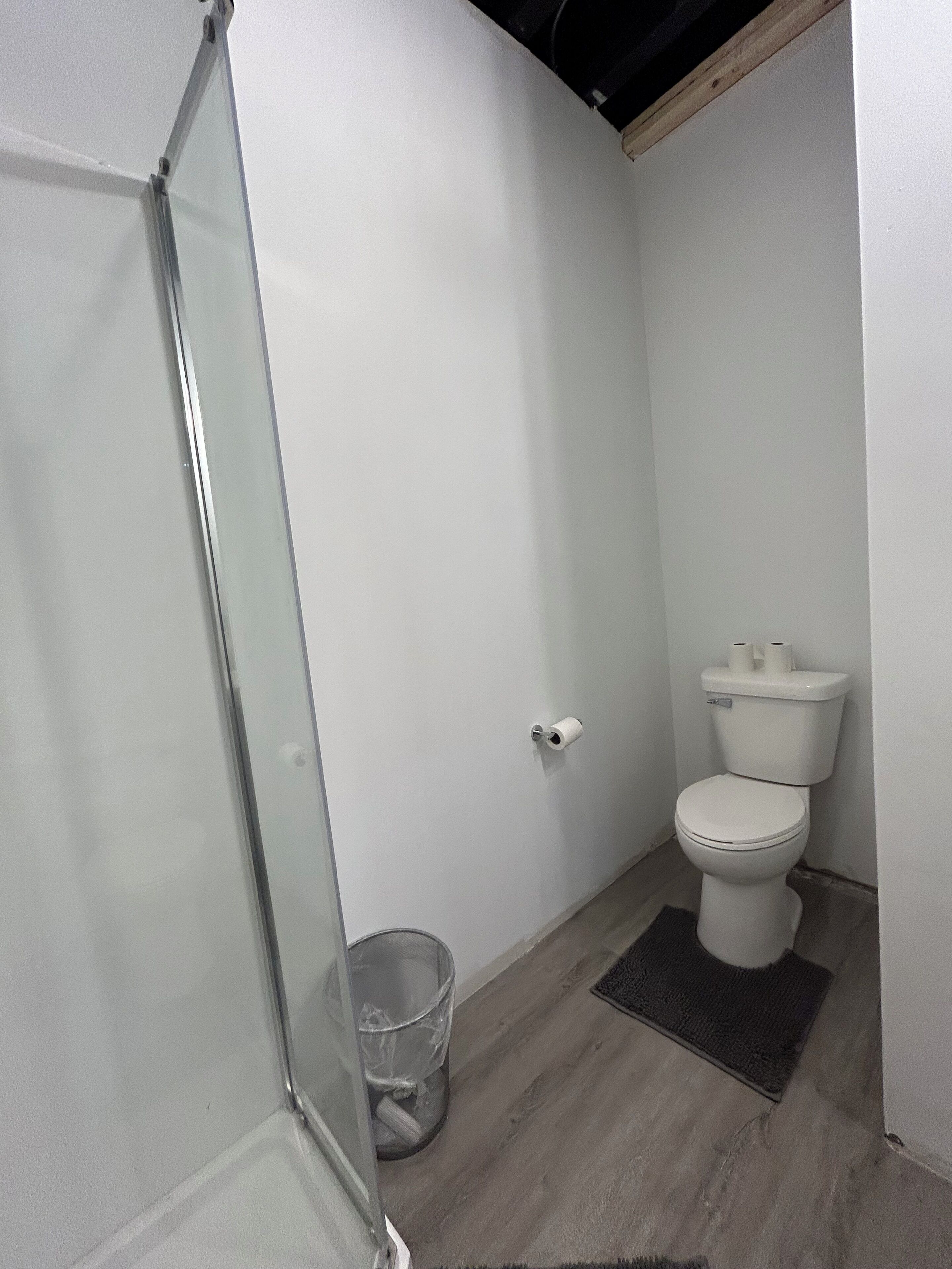 A small, modern bathroom with a glass shower enclosure, a toilet, and a trash bin.