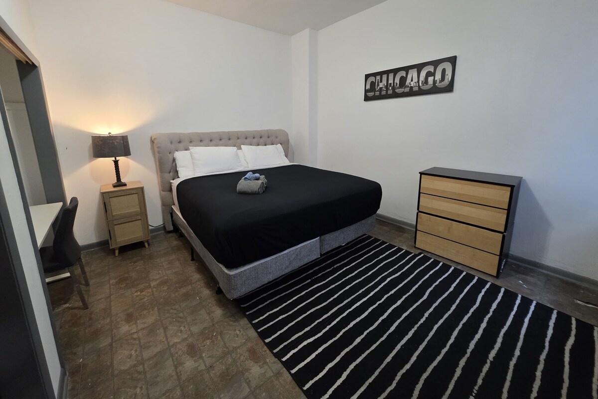 A bedroom with a bed, a nightstand, a dresser, a desk, and a wall sign.