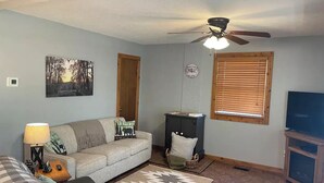 A living room with a sofa, a television, a ceiling fan, a clock, and a window with blinds. - A quiet retreat nestled right in the Hoosier National Forest. (Saint Croix)