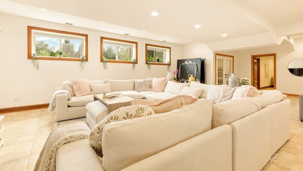 A spacious living room with a large L-shaped sectional sofa, three windows with plants, and a modern entertainment unit. - Luxury Suite in Glen Ellyn, IL USA (Glen Ellyn)