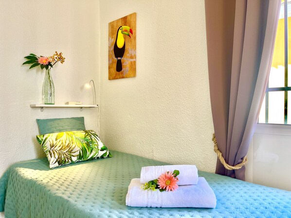 A neatly made bed with a green bedspread, a pillow with a tropical leaf pattern, a toucan painting on the wall, and a vase with flowers on a shelf.