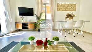 A modern living room with a glass coffee table, a flat-screen TV, a dining area with white chairs, and a stone wall decoration. - San Francisco Home by Gloove (Alicante / Alacant)