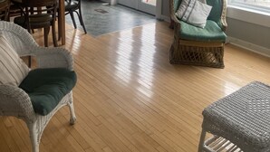 A spacious living room with a wooden floor, a dining area with a table and chairs, a wicker armchair, and a view through a glass door to the outside.