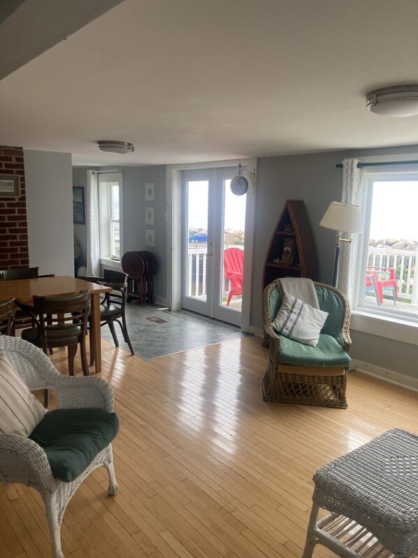 Un salón espacioso con piso de madera, un comedor con mesa y sillas, un sillón de mimbre, y vistas al exterior a través de un ventanal.