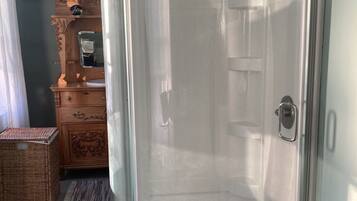 A glass-enclosed shower with a white floor, a wooden dresser with a mirror, and a wicker basket.