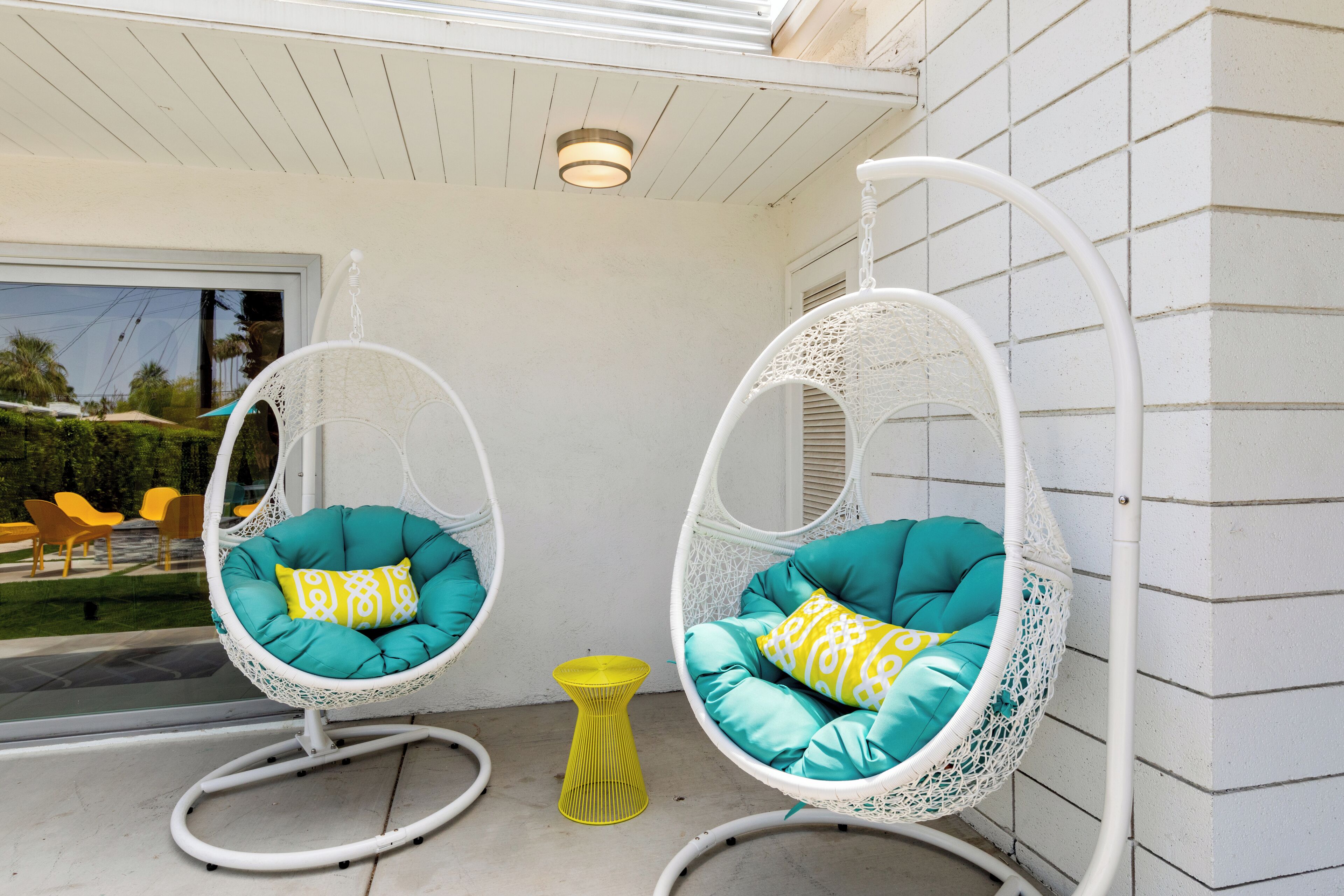 Two white wicker swing chairs with teal cushions and yellow patterned pillows, positioned on a patio with a yellow stool.