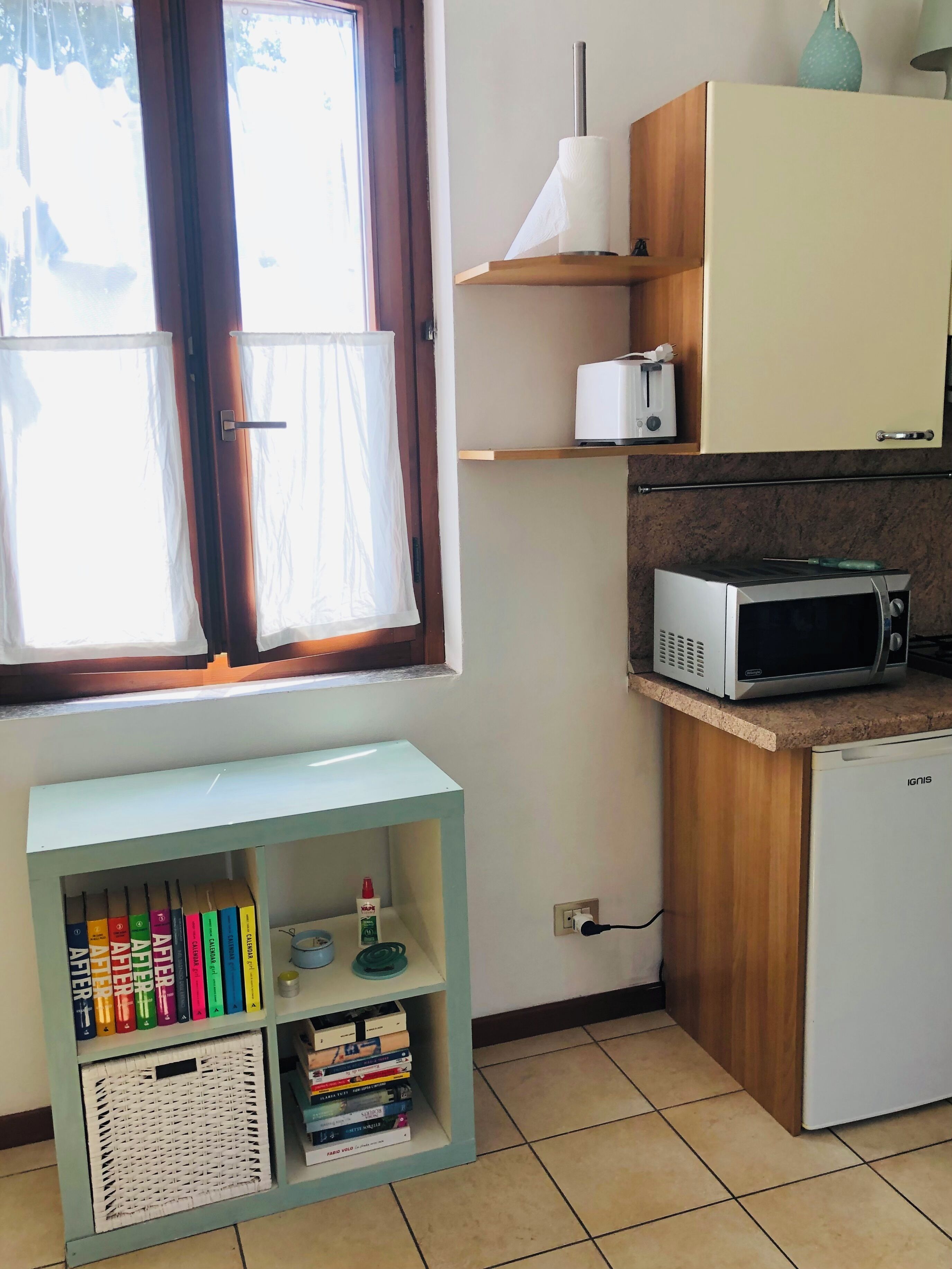 Une cuisine équipée d’un four à micro-ondes, d’un grille-pain et d’un petit réfrigérateur. Il y a une fenêtre avec un rideau, ainsi qu’une étagère avec des livres et une bouteille.