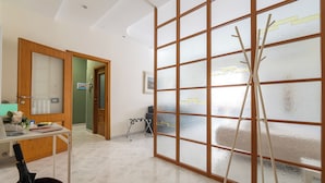 Habitación de hotel con puerta corrediza de vidrio, un escritorio con una computadora portátil, una silla, una mesita con un jarrón de flores y una escalera de madera apoyada en la puerta.