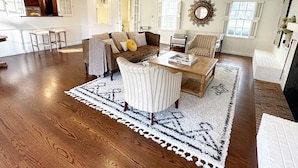 A spacious living room with a wooden floor, a sofa set, a coffee table, and a dining area with a kitchen in the background.