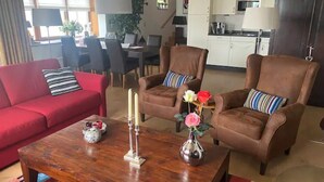 Una sala de estar con un sofá rojo, dos sillones marrones, una mesa de centro de madera y una cocina al fondo.