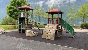 Un parque infantil con estructuras para escalar y toboganes, con montañas y un lago al fondo.