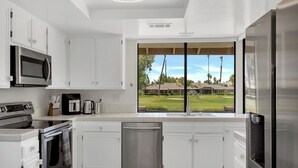 A modern kitchen with white cabinetry, stainless steel appliances, and a large window offering a view of a golf course. - Modern Desert Retreat (Palm Desert)