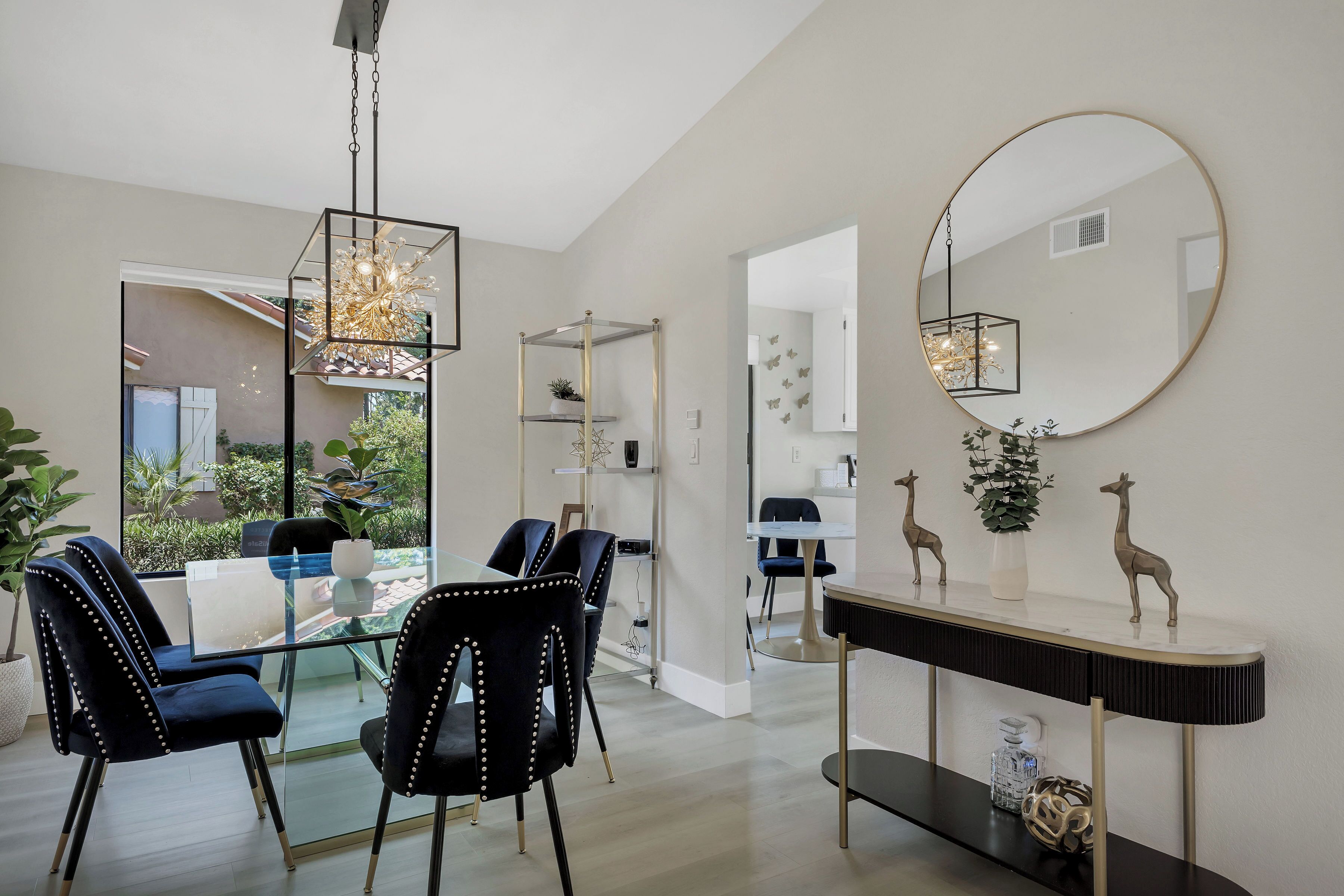 A modern dining area with glass tables and black upholstered chairs, a hanging light fixture, and decorative items on a console table.