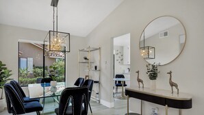 A modern dining area with glass tables and black upholstered chairs, a hanging light fixture, and decorative items on a console table. - Modern Desert Retreat (Palm Desert)