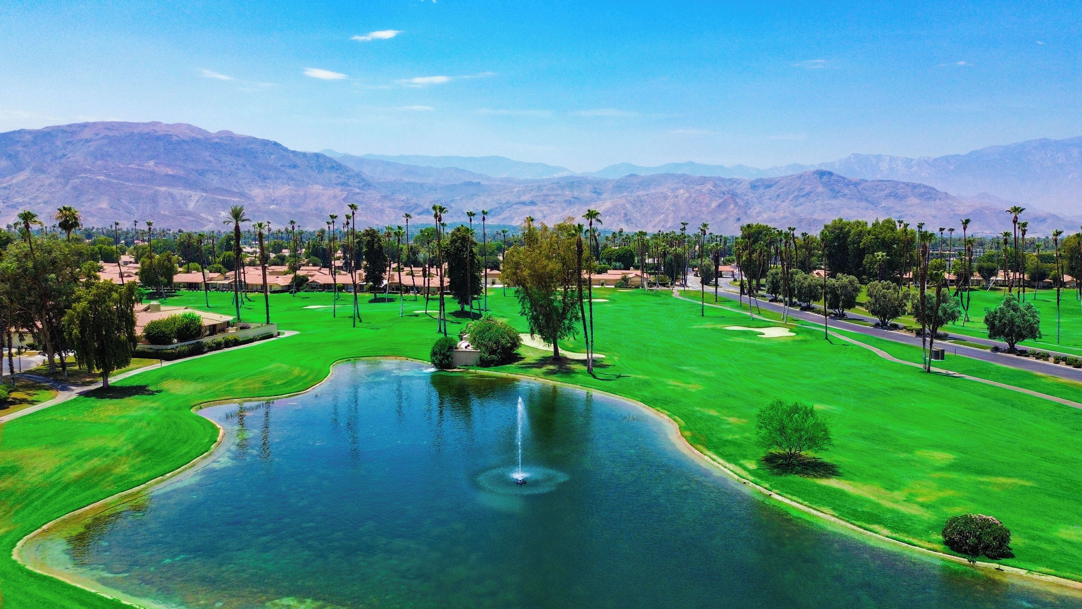 A golf course with a pond, palm trees, and mountains in the background.