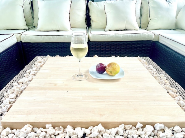 A wooden table with a glass of white wine and two fruits on a plate, surrounded by pebbles, with a wicker sofa and white cushions in the background.