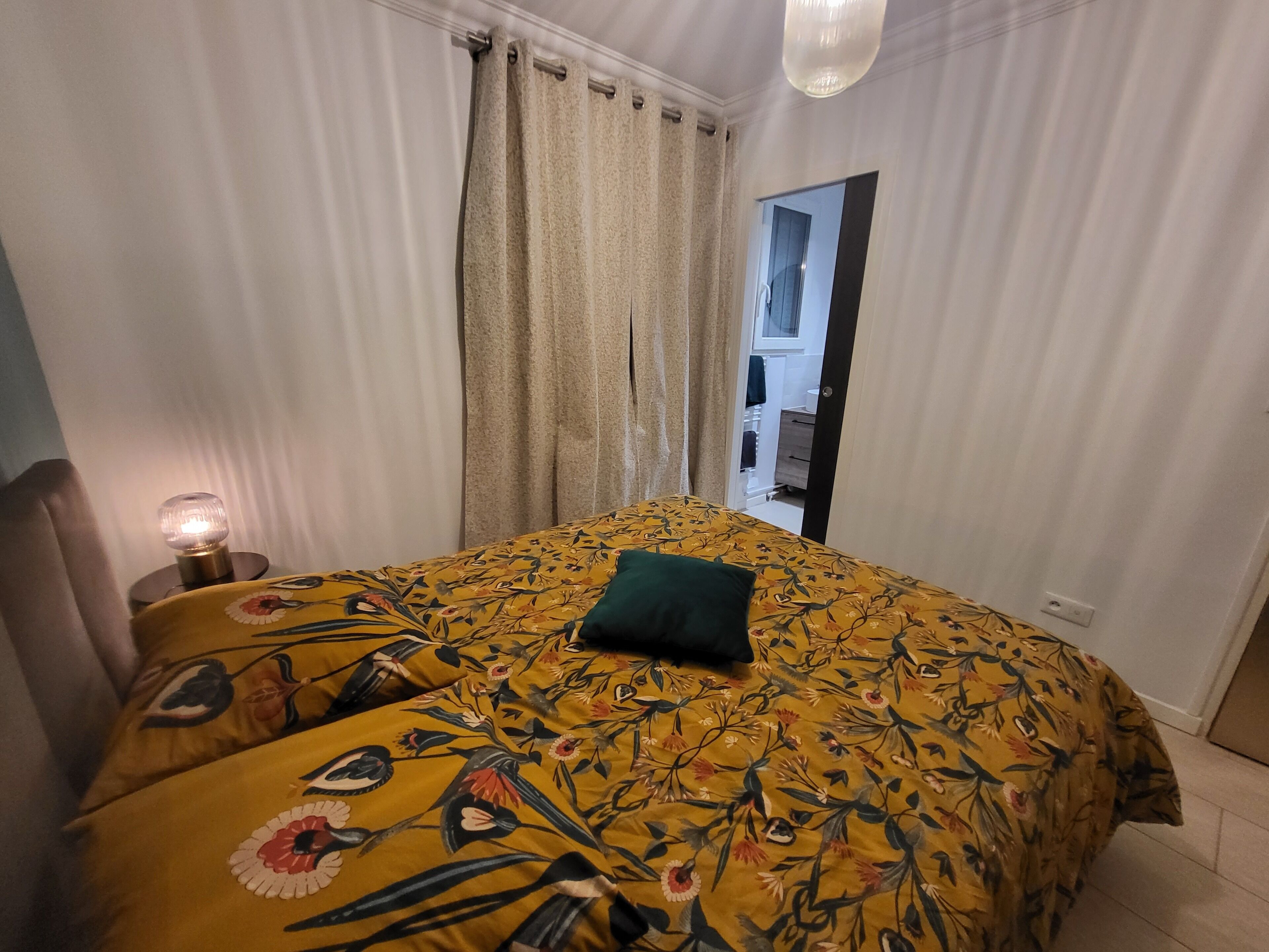 Un dormitori amb un llençol floral, un coixí verd, una làmpada de nit i vista a un bany per una porta oberta.