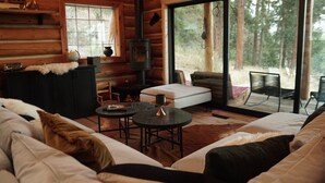 A cozy cabin interior with a sofa, a small table, a fireplace, and a view of the forest through large windows. - Teanaway Pines: Riverfront Mountain Retreat (Cle Elum)