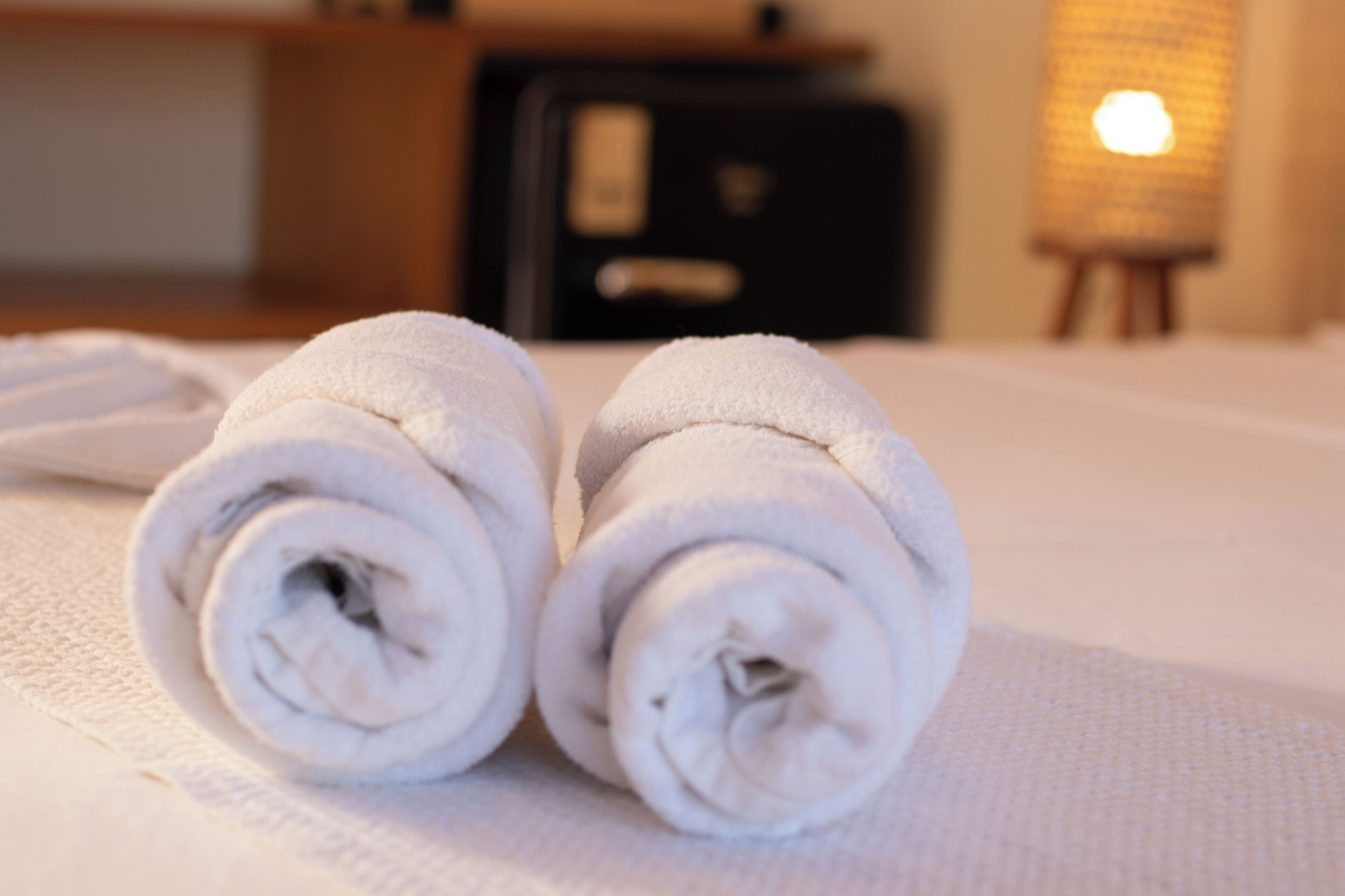 Dos toallas blancas enrolladas sobre una superficie texturizada, posiblemente una cama o encimera, con un fondo desenfocado que incluye una lámpara y un mueble.