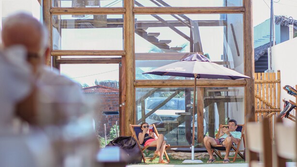 Una estructura de fusta amb teulada de palla, tancament de vidre i zona de piscina amb gandules.