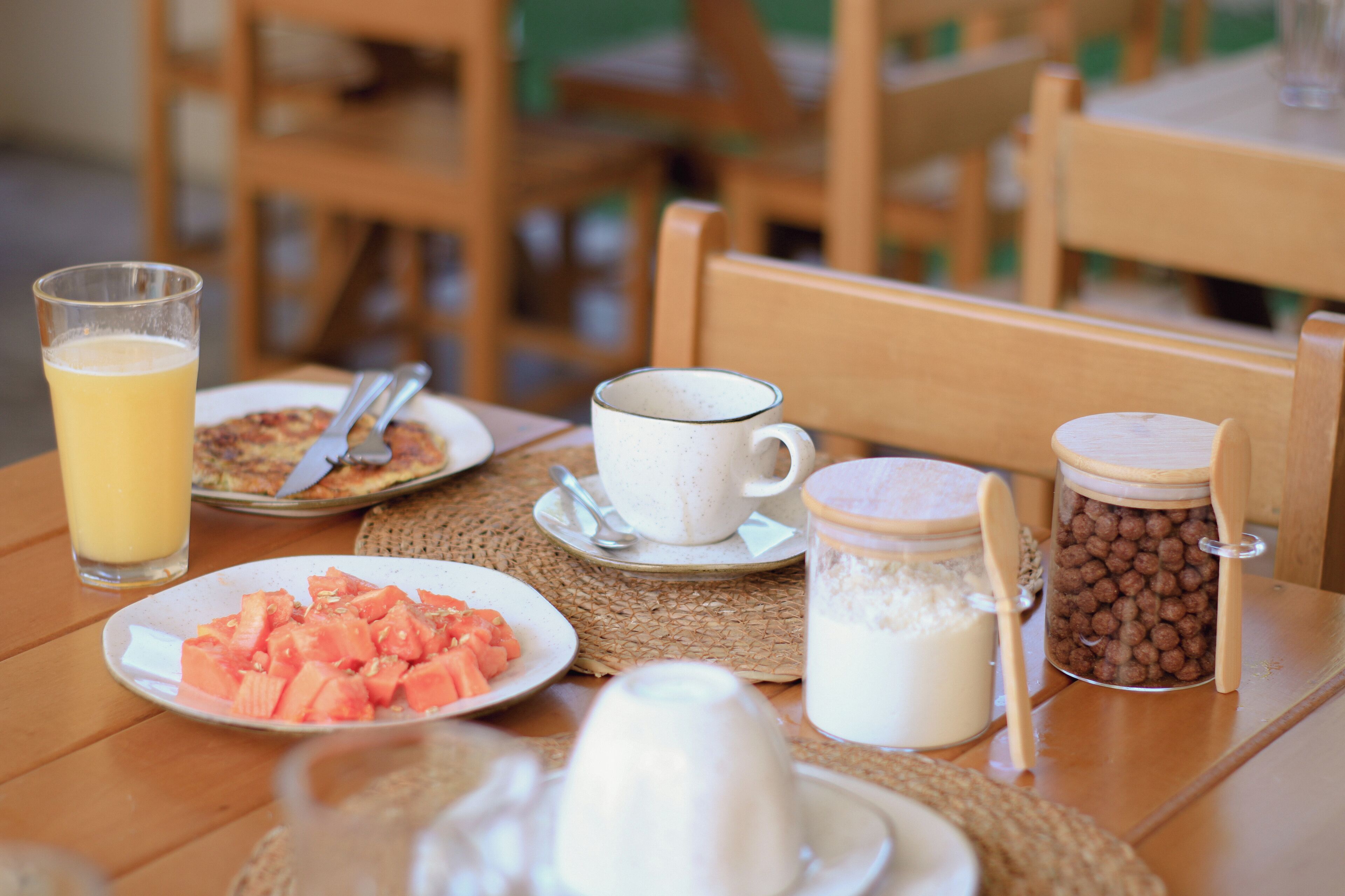 A breakfast table with a glass of orange juice, a plate of watermelon, a cup of coffee, a jar of chocolate balls, and a jug of milk.