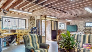 A spacious room with exposed wooden beams, a stone wall, and a large window. There are two upholstered chairs, a potted plant, and a desk with a computer and a Renault truck oil sign. - Cozy home in Neuilly le Vendin with WiFi (Neuilly le Vendin)