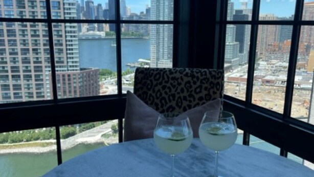 A table with two glasses of a light-colored beverage, a leopard print pillow, and a view of a cityscape and water through large windows. - Gorgeous apt for Labor Day weekend.  View of Manhattan at Greenpoint 



 (Brooklyn)