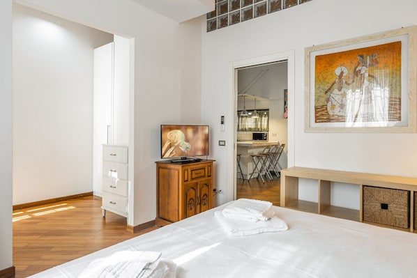 A bedroom with a bed, a TV, a wooden cabinet, a painting, and a kitchenette in the background.