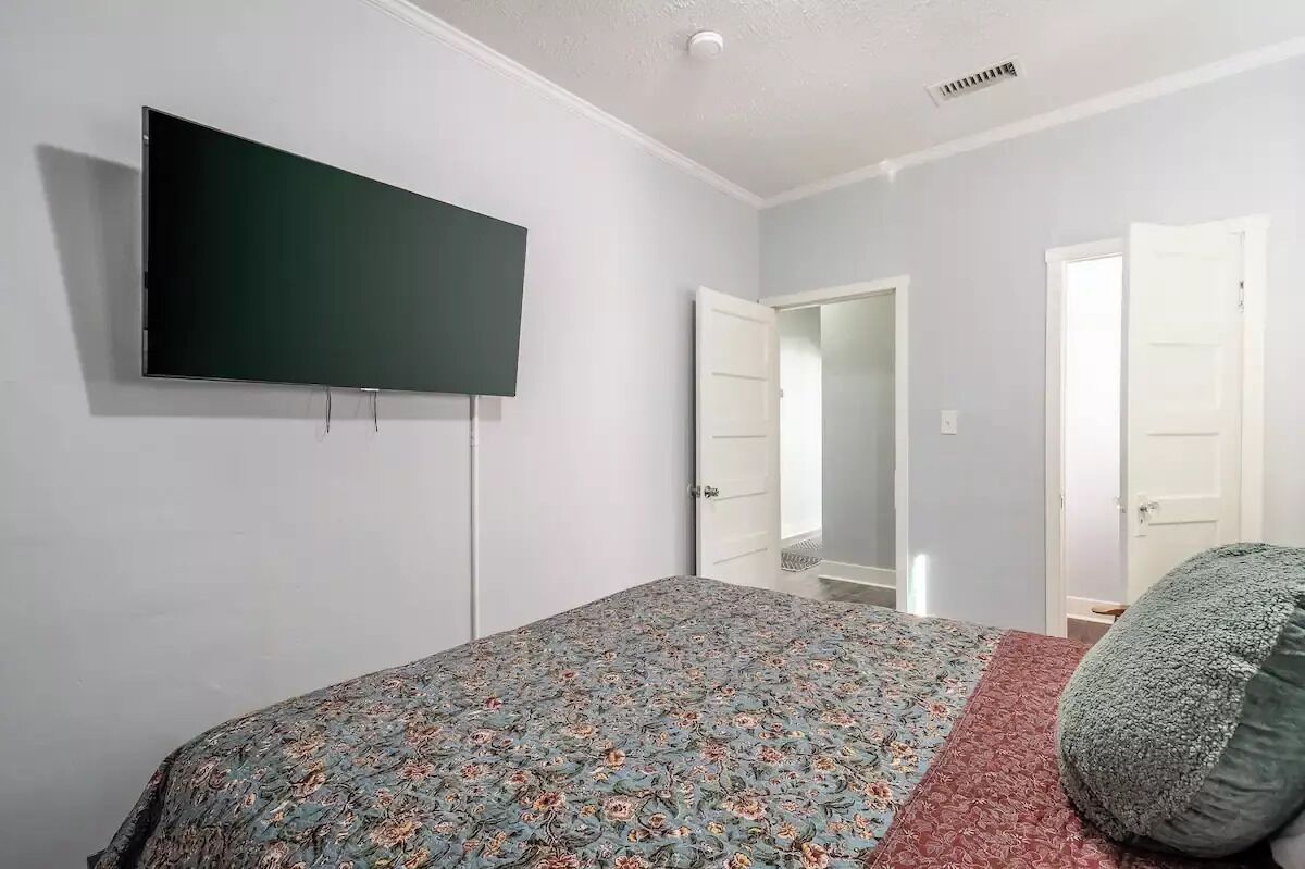 A bedroom with a bed, a flat-screen TV mounted on the wall, and a door leading to another room.