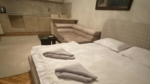 A bedroom with a bed, a sofa, a painting on the wall, and a kitchen area in the background. - APARTMENT PERLA (Pristina)