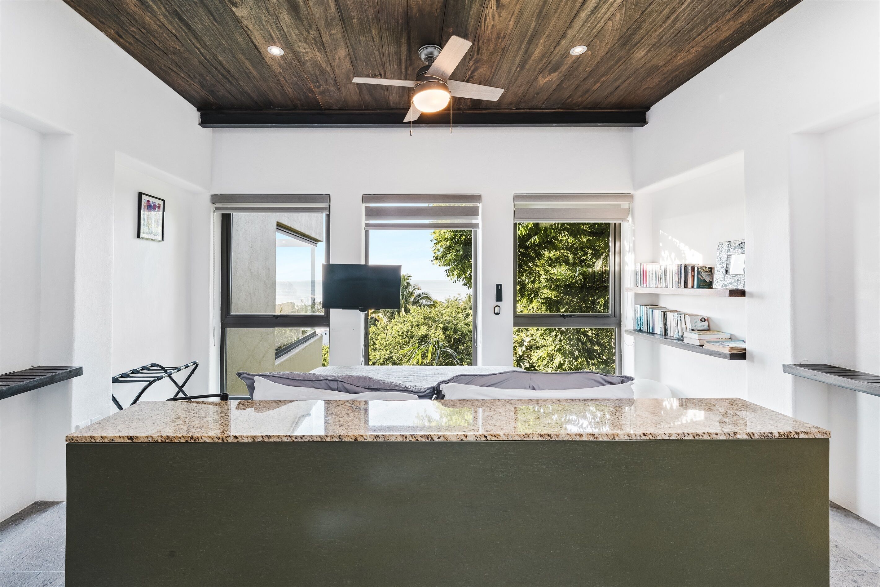 A modern kitchen with a large island, wooden ceiling, and large windows offering views of greenery.