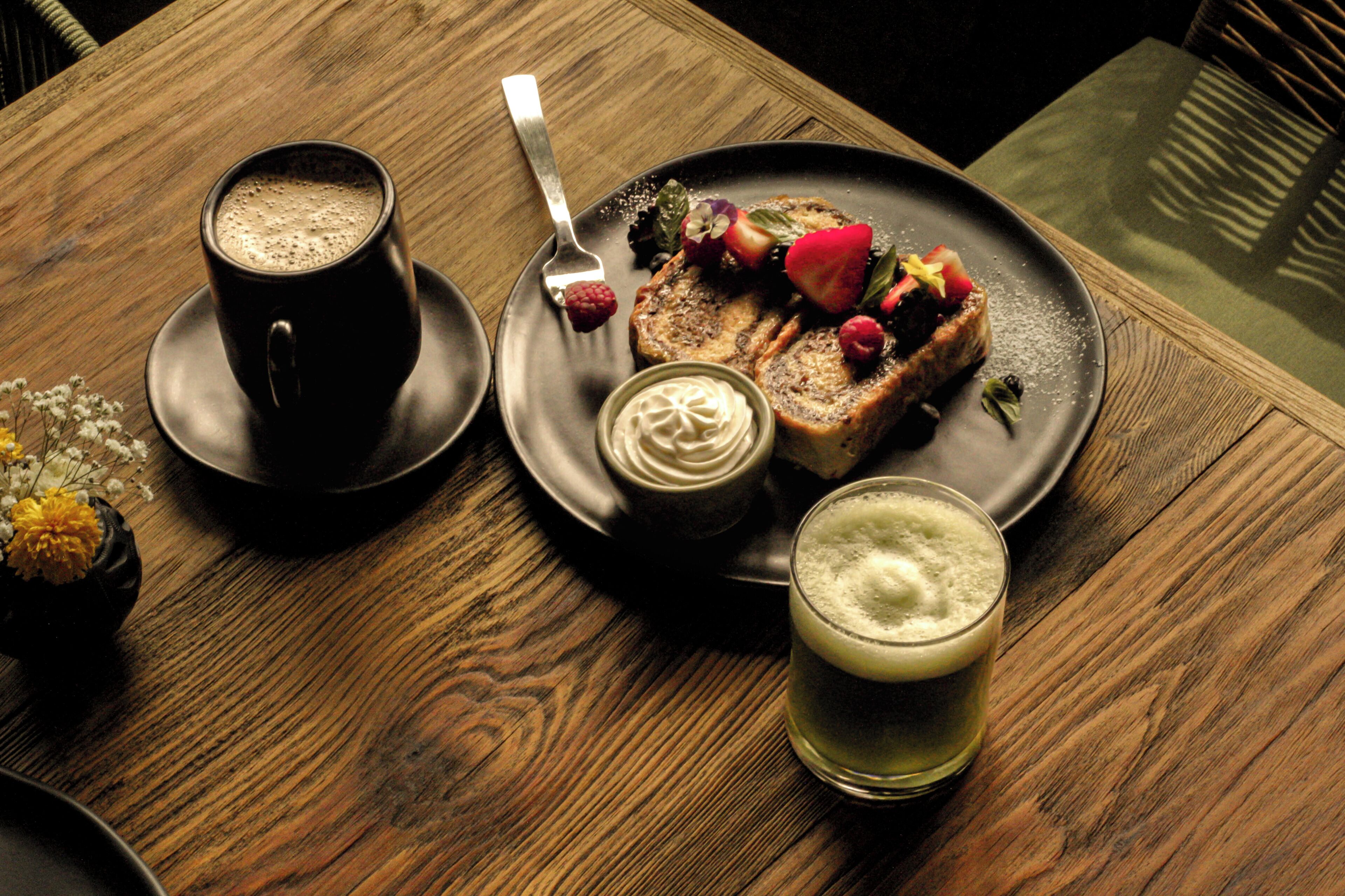 Une table en bois sur laquelle se trouvent une assiette de nourriture, une tasse de café et un verre contenant une boisson de couleur claire.