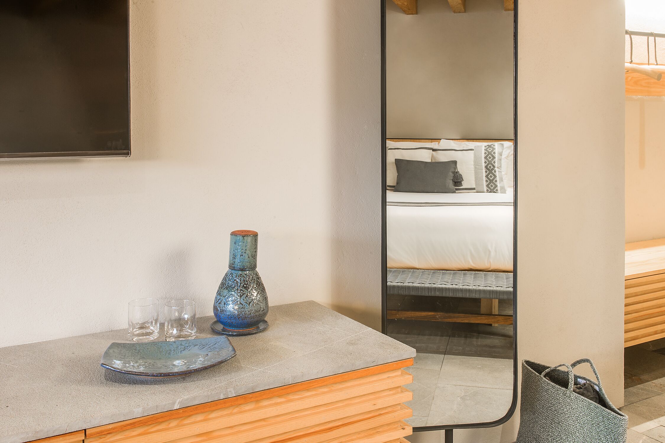 Habitación de hotel con una cama, una mesita de noche con un jarrón y vasos, y un espejo que refleja la cama y una ventana.
