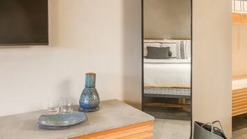 Habitación de hotel con una cama, una mesita de noche con un jarrón y vasos, y un espejo que refleja la cama y una ventana.