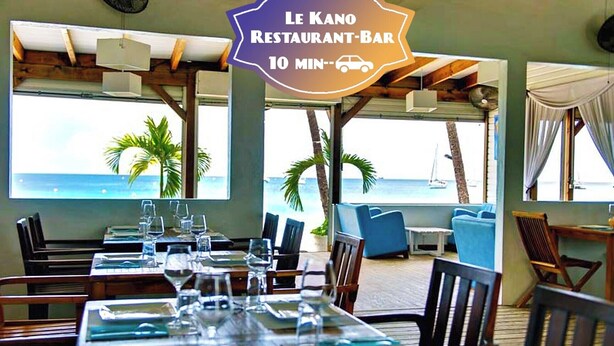 A restaurant-bar with a view of the ocean, tables set with glasses and plates, and a sign indicating a 10-minute car service. - Vue mer Imprenable ! F2 2/4 Pers, Jacuzzi Privatif et Piscine Privée (Les Trois-Îlets)