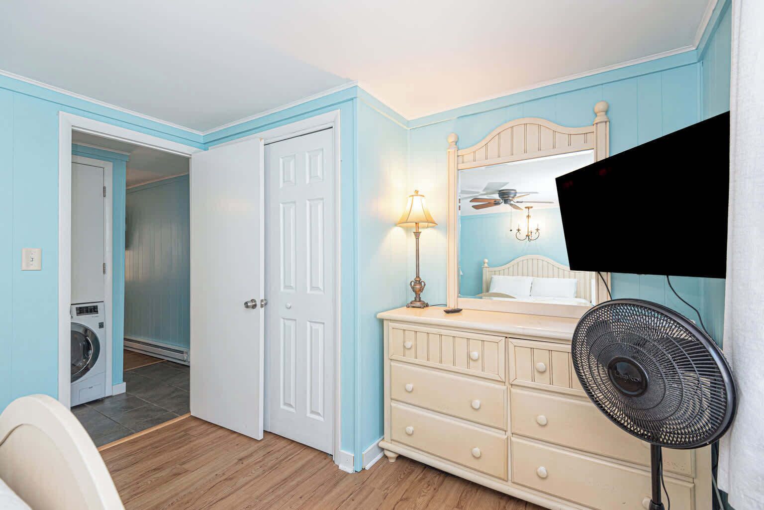 Quarto com cama, cômoda, espelho, luminária, ventilador de teto e máquina de lavar no cômodo vizinho.
