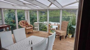 Une véranda avec une table et des chaises, un fauteuil en osier, une petite table avec une étagère et des plantes en pot.