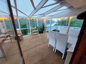 A spacious deck with wooden flooring, white cushioned seating, and a glass ceiling offering a view of the outdoors. - Cozy village house in Funningsfjørð (Funingsfjord)