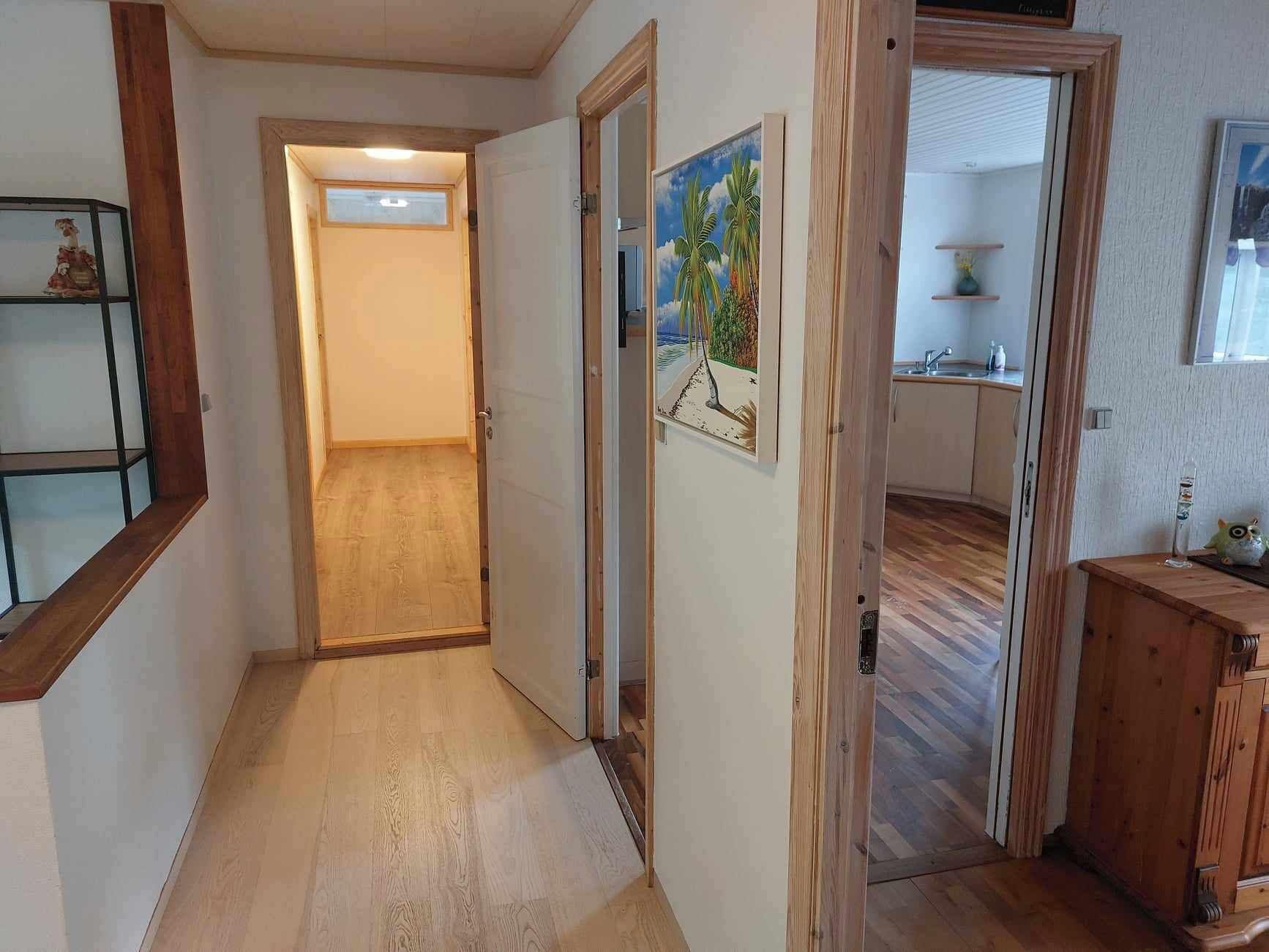 Un couloir doté d’un plancher en bois, une porte donnant sur une pièce où se trouve un tableau, et un meuble en bois.