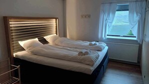Un lit bien fait, avec du linge de lit blanc et des serviettes pliées, une tête de lit en bois et une fenêtre avec des rideaux.