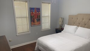 Un dormitorio con una cama grande, dos ventanas con persianas, una mesita de noche con lámpara y un cartel en la pared.
