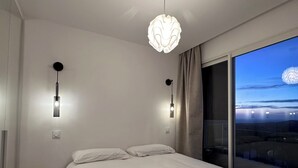 A modern bedroom with a large bed, two wall-mounted lights, and a view of the city through a large window.