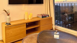 A modern living room with a flat-screen TV mounted on the wall, a wooden TV stand with books and candles, a small round table with a perfume bottle, and a large window with a view of the cityscape.